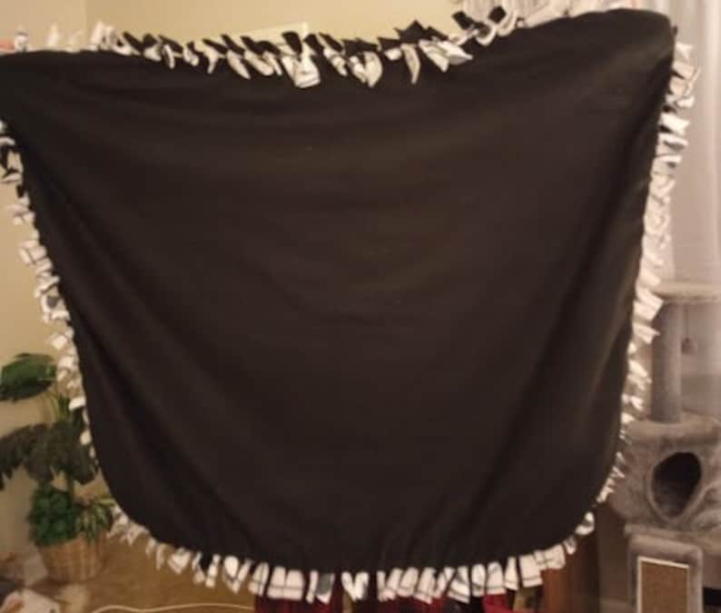 Puede incluir: Una manta de forro polar negra con un borde de flecos anudados blancos. La manta est&aacute; doblada por la mitad y puesta plana sobre una superficie.