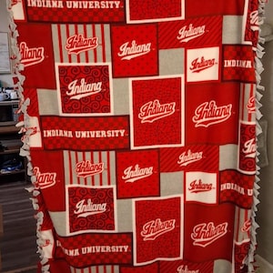 Op de afbeelding: Een fleece deken met een patchwork ontwerp van rode en witte vierkanten met Indiana University branding. Het patroon bevat verschillende texturen zoals ruiten, strepen en krullen, die allemaal het Indiana-logo tonen.