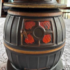 May include: Black ceramic cookie jar shaped like a wood-burning stove with a red and black painted design. The jar has a lid and a gold trim.