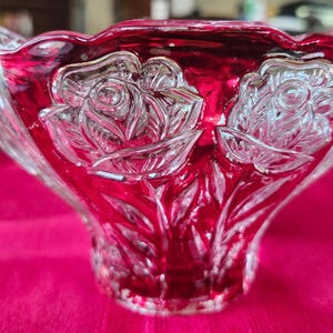 May include: A clear glass bowl with a ruby red interior and rim. The bowl features embossed rose designs. The bowl is sitting on a bright pink surface. The bowl is likely used for decorative purposes.