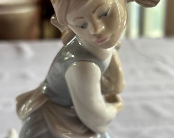 May include: A porcelain figurine of a young girl with blonde hair in pigtails, wearing a light blue dress. The girl is holding a small object, with her eyes closed. The figurine has a glossy finish.