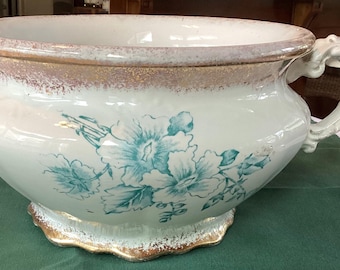 May include: Antique white porcelain planter with a wide rim and ornate handle. The planter features a floral design in shades of teal and gold accents. A decorative piece for indoor plants or as a vintage decor accent.