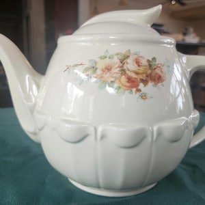 May include: Cream-colored ceramic teapot with a floral rose design. The teapot has a curved spout, a handle, and a lid with a decorative finial. The body of the teapot features a scalloped design along the bottom.
