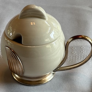 May include: Cream-colored ceramic sugar bowl with a lid and a silver handle. The bowl has a round shape with a silver accent on the side. The handle is a curved, silver metal. The bowl sits on a silver base.