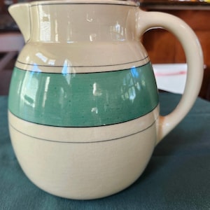 May include: Cream-colored ceramic pitcher with a rounded body and a curved handle. The pitcher features a wide teal band with black trim, and a spout for pouring. The pitcher is a vintage style and is suitable for serving beverages.