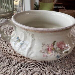 May include: A large, white ceramic bowl with a floral design. The bowl has a wide, round opening and a single handle. The floral design features pink and blue flowers with green leaves.