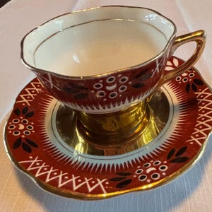 May include: A vintage teacup and saucer set. The teacup has a white interior and a maroon exterior with a floral design. The saucer is maroon with a gold rim and a matching floral pattern. The handle and base are gold.