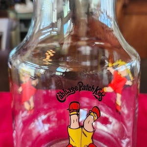 May include: Clear glass pitcher with a wide base and a flared top. The pitcher is decorated with colorful Cabbage Patch Kids graphics. The Cabbage Patch Kids logo is also visible. The pitcher is likely vintage and collectible.