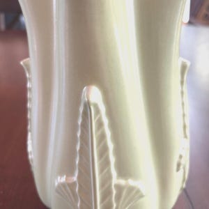 May include: A cream-colored ceramic vase with a fluted design. The vase features a repeating leaf pattern and a flared top. The vase is approximately 10 inches tall and would be a decorative addition to any home.