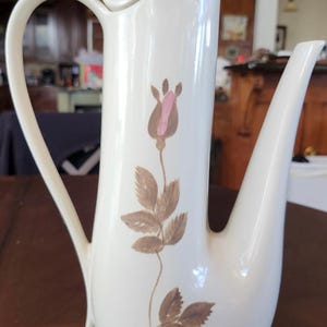 May include: Cream-colored ceramic coffee pot with a tall, slender design. It features a curved handle and a spout. A floral design with a pink bud and brown leaves decorates the front. The pot is likely vintage.