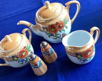 May include: A vintage tea set with a teapot, sugar bowl, creamer, and two salt shakers. The set features a peach-colored glaze with floral and butterfly designs in orange, green, and blue. The set is on a blue surface.