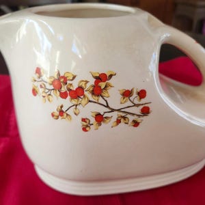 May include: Cream-colored ceramic pitcher with a wide handle and a decorative floral design. The pitcher features a branch with red berries and yellow leaves. The pitcher has a wide opening and a rounded base, suitable for serving beverages.