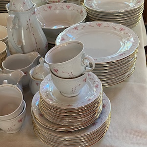 May include: A collection of white porcelain dinnerware with a delicate pink floral pattern and gold trim. The set includes plates, cups, saucers, a teapot, serving bowls, and a sugar bowl, all arranged on a white tablecloth. The tableware is suitable for a formal dining setting.