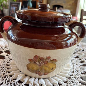 May include: A brown and white ceramic crock with a lid. The crock has a design of two apples and leaves on the front. The crock is sitting on a white crocheted doily.