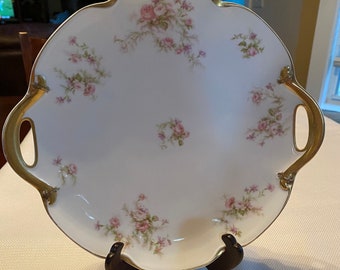 May include: A white porcelain serving platter with a scalloped edge and two gold handles. The platter is decorated with a delicate floral pattern of pink roses and green leaves.