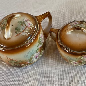 May include: A vintage brown and white teapot and creamer set with floral designs. The teapot has a lid and a handle. The creamer has a lid and a handle.