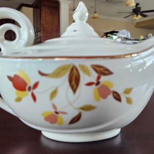 May include: White ceramic teapot with a curved handle and spout. The teapot features a floral design in red, yellow, and brown, with gold trim. The lid has a decorative finial. A vintage kitchenware item.