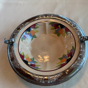 May include: A vintage ceramic bowl with a floral design in shades of blue, red, yellow, and green. The bowl is surrounded by a silver-plated rim with a decorative pattern.