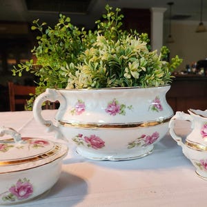 May include: A white porcelain serving set with pink rose floral designs and gold trim. The set includes a large handled planter with greenery, a small creamer, and a lidded bowl. The pieces are arranged on a white tablecloth.