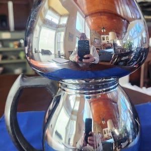 May include: A vintage chrome coffee pot with a black handle and lid. The pot has a reflective surface, showing a distorted reflection of the surroundings. The pot sits on a blue cloth.