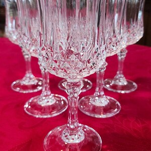 May include: A set of six clear crystal wine glasses. Each glass features a diamond-cut pattern on the bowl and a long, elegant stem. The glasses are arranged on a red tablecloth, showcasing their intricate design and clarity.