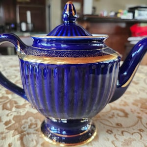 May include: A cobalt blue ceramic teapot with a gold trim. The teapot has a ribbed design and a curved handle. The lid is topped with a gold finial. A classic piece for tea parties or display.