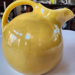May include: A yellow ceramic pitcher with a rounded body, a curved handle, and a spout. The pitcher has a glossy finish and reflects the surrounding environment. The pitcher is likely used for serving beverages.