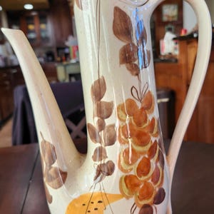May include: Tall, cream-colored ceramic pitcher with a long spout and curved handle. The pitcher is decorated with brown and orange floral designs. The pitcher is sitting on a brown table.