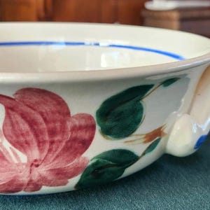 May include: A white ceramic bowl with a handle, decorated with a floral pattern in red, yellow, and green. The handle is white with blue accents, and a blue stripe encircles the rim. A decorative kitchenware item.