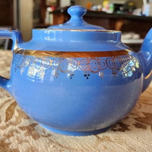 May include: A blue ceramic teapot with a lid, handle, and spout. The teapot is decorated with a gold band and gold filigree designs. The teapot is sitting on a patterned tablecloth.