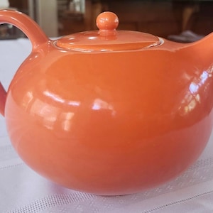 May include: A glossy, orange ceramic teapot with a rounded body, curved handle, and a spout. The lid has a small knob. The teapot is sitting on a white tablecloth. This teapot is perfect for serving tea.