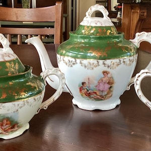 May include: A porcelain tea set with a teapot, sugar bowl, and creamer. The set features a white base with green and gold accents. Each piece has a decorative handle and a painted scene.