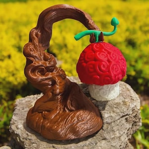 May include: A brown, stylized tree sculpture with a red, textured fruit and a white base. The fruit has a green stem and is set on a rock. The sculpture is outdoors with a blurred green and yellow background.