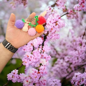 May include: A keychain with five colorful charms shaped like fruit. The charms are purple, white, pink, orange, and red. The keychain is hanging from a hand against a background of pink cherry blossoms.
