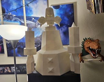 May include: A large, off-white, geometric sculpture with a central tower and two flanking structures. The sculpture has a futuristic design, with sharp angles and triangular accents. A floor lamp with a white shade is to the left. A dinosaur head is on the right.