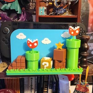 May include: A colorful 3D-printed Super Mario Bros. themed display. Features green pipes, brick blocks, a question mark block, a star, and Piranha Plants against a blue backdrop with white clouds. The display is placed in front of a black game console.