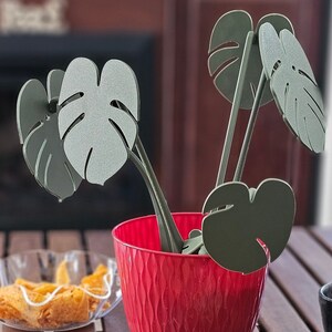 May include: A red planter with a textured surface holds a set of green, leaf-shaped decorations. The decorations are made of a lightweight material and resemble a tropical plant.