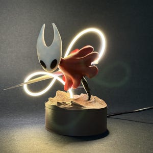 May include: A detailed figurine of a character with a white head, red cape, and a long, thin blade. The figure is illuminated by a glowing neon light, mounted on a small, textured base. A black power cord extends from the base.