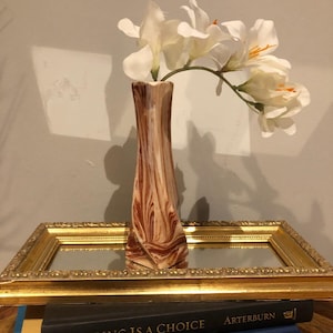 May include: A tall, slender vase with a brown and cream marbled design holds white flowers with orange centers. The vase sits on a gold-framed mirror, with books visible below. The book on top reads "HEALING IS A CHOICE".
