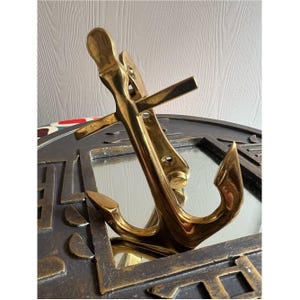 Op de afbeelding: Een messing anker, een nautisch decoratief item, is het middelpunt. Het anker is schuin geplaatst, met de vloeien en de schacht zichtbaar. Het anker staat tegen een donkere, mogelijk metalen achtergrond met geometrische ontwerpen.
