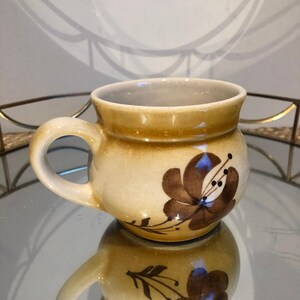 May include: A ceramic mug with a rounded body and a handle. The mug is beige with a brown floral design. The mug is reflected in a mirrored surface.