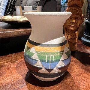 Può includere: Un vaso in ceramica bianca con un motivo geometrico in tonalità di verde, blu, giallo e nero. Il vaso ha una base arrotondata e un'ampia apertura.