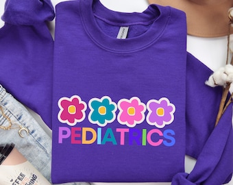 May include: Purple sweatshirt with a rainbow colored word "PEDIATRICS" and five colorful flower designs.