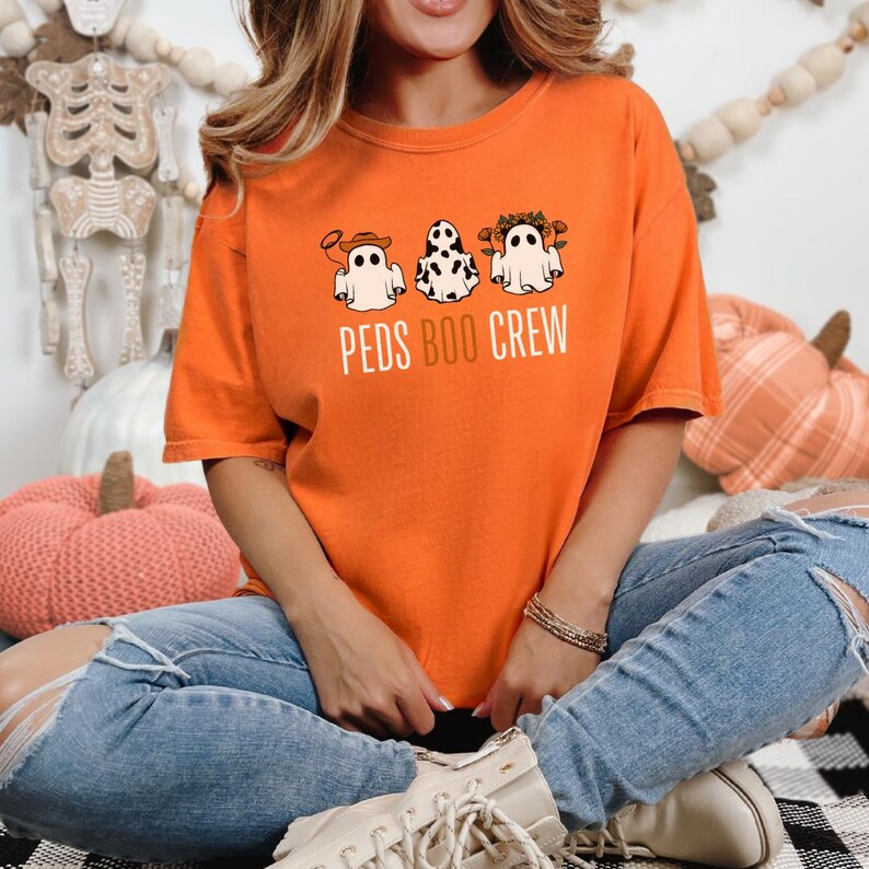Puede incluir: Camiseta naranja con un gr&aacute;fico de tres fantasmas con diferentes disfraces. El texto "PEDS BOO CREW" est&aacute; impreso debajo de los fantasmas.