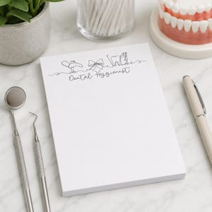 May include: A white notepad with the words "Dental Hygienist" and a line drawing of dental tools. A dental mirror and probe are on the left. A pen and a model of teeth are on the right. A small plant and cotton swabs are in the background.
