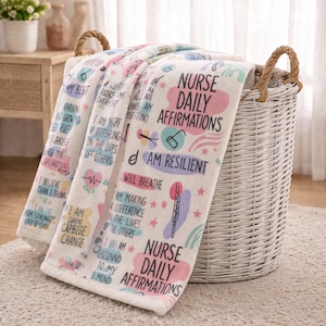 May include: A white fleece blanket with the text "Nurse Daily Affirmations" in pink and blue. The blanket has various affirmations printed on it, and is folded over a white wicker basket with brown handles.