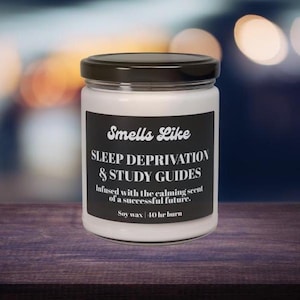 May include: A white candle in a glass jar with a black lid. The black label reads "Smells Like Sleep Deprivation & Study Guides." The candle is made from soy wax, infused with a calming scent, and has a 40-hour burn time. The jar is on a wooden surface.