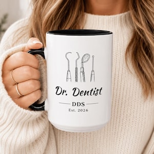 May include: A white ceramic mug with a black interior and handle, featuring a black line art illustration of four dental tools above the text Dr. Dentist, DDS, and Est. 2026 printed in a black serif and script font on the front surface.