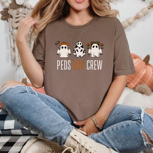 Puede incluir: Una camiseta marr&oacute;n con un gr&aacute;fico de tres fantasmas que llevan diferentes accesorios. El texto "PEDS BOO CREW" est&aacute; impreso debajo de los fantasmas.