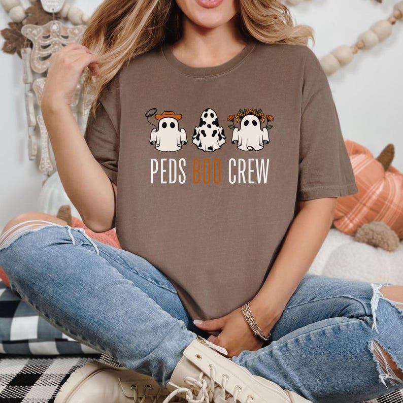 Puede incluir: Una camiseta marr&oacute;n con un gr&aacute;fico de tres fantasmas que llevan diferentes accesorios. El texto "PEDS BOO CREW" est&aacute; impreso debajo de los fantasmas.
